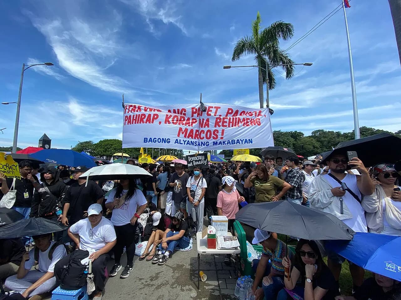 Protesters under umbrellas behind a banner against corruption and the Marcos administration